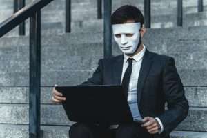 Businessman with mask on his face looks into the laptop. Sits on the steps of a business building