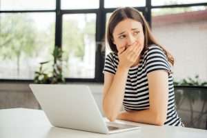 sad girl in striped t-shirt covering mouth with hand while using laptop at home