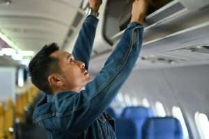 Shot of hipster man passengers putting his luggage into the overhead compartment on an airplane.