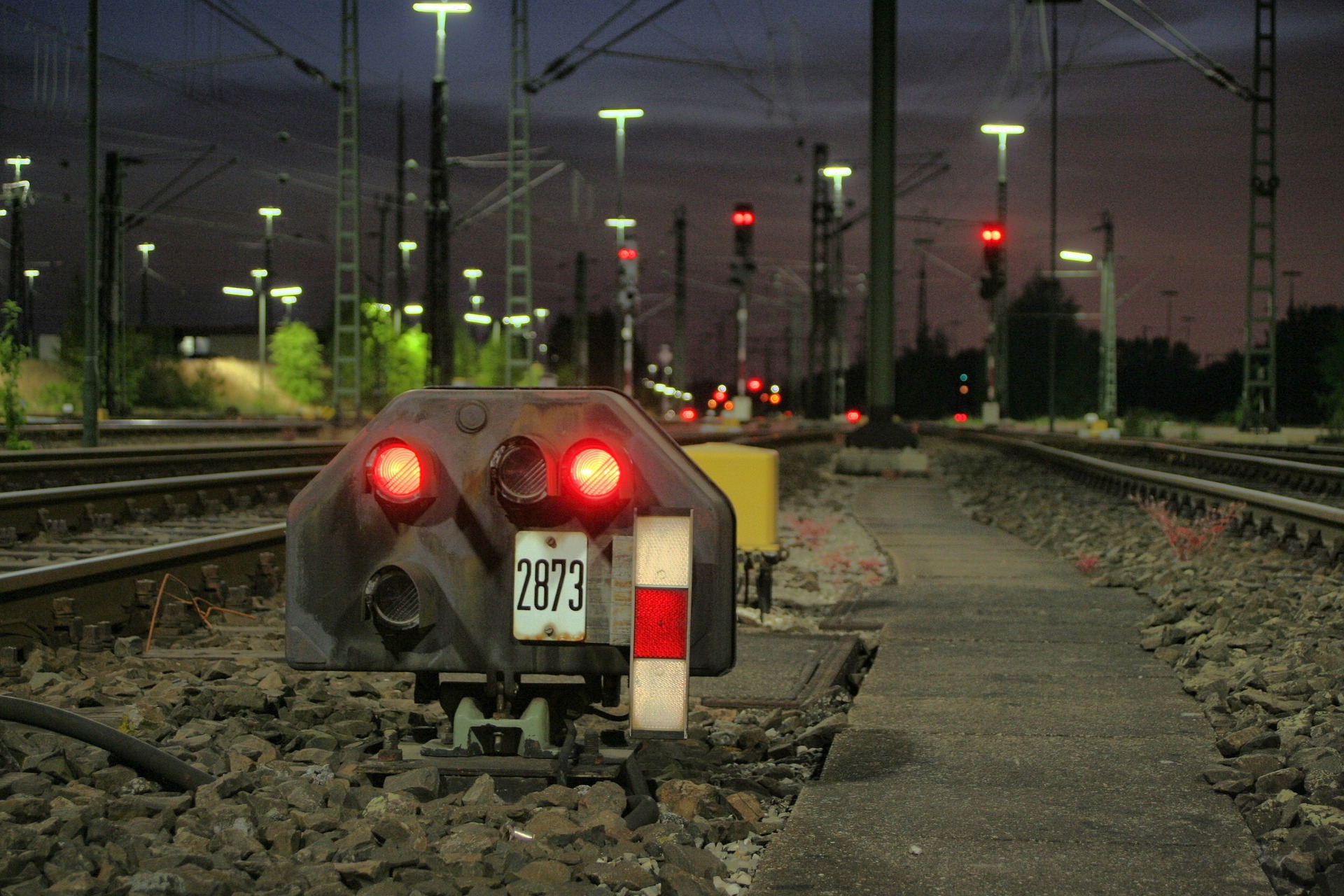 A red railway stop signal beside the tracks in a rail yard at dusk, with the lines running into the distance